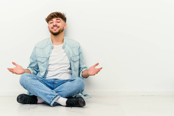 Faslı genç bir adam beyaz arka planda izole bir şekilde yerde oturuyor rahat ve mutlu bir şekilde gülüyor, boynunu uzatmış dişlerini gösteriyor. - Fotoğraf, Görsel