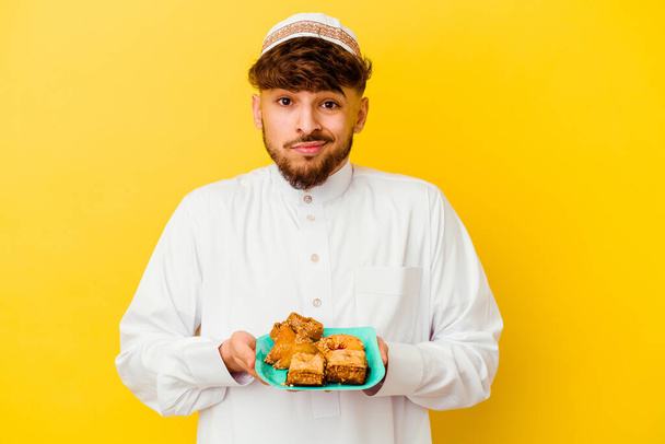 Νεαρός Μαροκινός άνδρας φορώντας το τυπικό αραβικό κοστούμι τρώει αραβικά γλυκά που απομονώνονται σε κίτρινο φόντο - Φωτογραφία, εικόνα