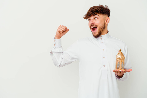 Jeune homme marocain portant un vêtement arabe typique tenant une lampe de ramadan isolé sur fond blanc levant le poing après une victoire, concept gagnant. - Photo, image