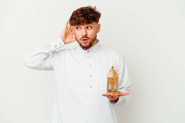 Junger marokkanischer Mann in typischer arabischer Kleidung, der eine Ramadan-Lampe auf weißem Hintergrund hält und versucht, Klatsch und Tratsch zu hören. - Foto, Bild