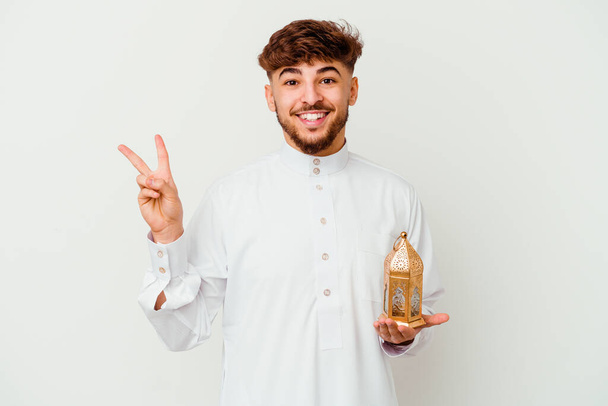 Młody Marokańczyk w typowym arabskim ubraniu, trzymający lampę ramadanu na białym tle, radosny i beztroski, pokazujący symbol pokoju palcami.. - Zdjęcie, obraz