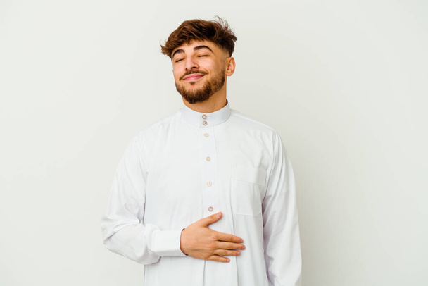Młody Marokańczyk noszący typowe arabskie ubrania odizolowane na białym tle dotyka brzucha, uśmiecha się delikatnie, jedząc i satysfakcjonując koncepcję. - Zdjęcie, obraz