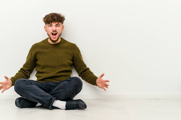 Joven hombre marroquí sentado en el suelo aislado sobre fondo blanco gritando muy enojado, concepto de rabia, frustrado. - Foto, imagen