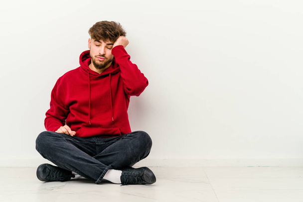 Jovem marroquino sentado no chão isolado no fundo branco cansado e muito sonolento mantendo a mão na cabeça. - Foto, Imagem