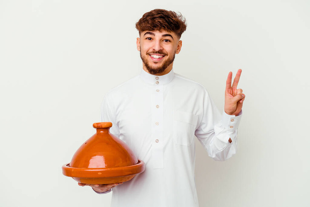 Junger marokkanischer Mann im typischen arabischen Kostüm mit einer Tajine auf weißem Hintergrund, fröhlich und unbeschwert, die ein Friedenssymbol mit den Fingern zeigt. - Foto, Bild