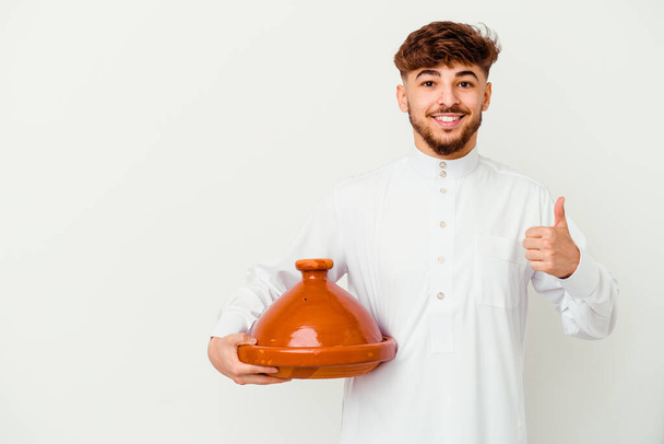 Mladý marocký muž v typickém arabském kostýmu drží tajine izolované na bílém pozadí s úsměvem a zvedá palec nahoru - Fotografie, Obrázek