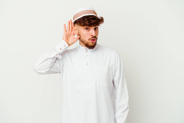 Junger arabischer Mann im typischen arabischen Kostüm, isoliert auf weißem Hintergrund, versucht einen Klatsch und Tratsch zu hören. - Foto, Bild