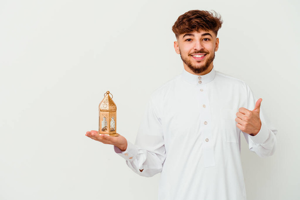 Junger marokkanischer Mann in typischer arabischer Kleidung hält eine Ramadan-Lampe auf weißem Hintergrund, lächelt und hebt den Daumen - Foto, Bild