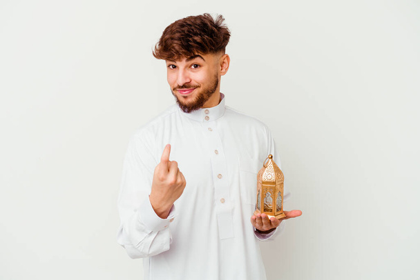 Joven marroquí vestido con una típica ropa árabe sosteniendo una lámpara de ramadán aislada sobre fondo blanco señalándote con el dedo como si invitaras a acercarte. - Foto, Imagen
