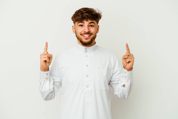 Beyaz arka planda izole edilmiş tipik Arap giysileri giyen genç Faslı bir adam iki ön parmağı da boş bir alan göstermekte.. - Fotoğraf, Görsel