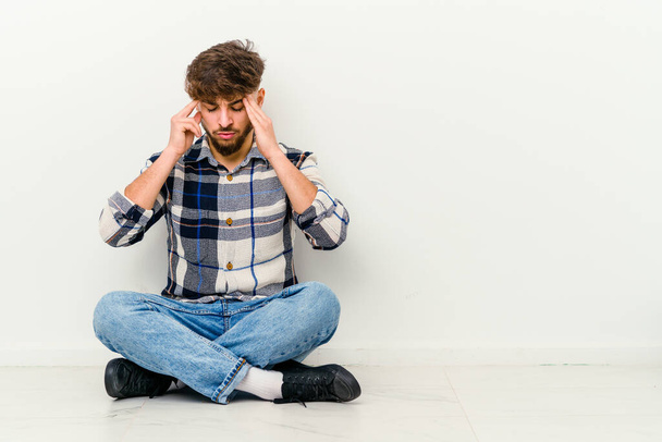 Joven hombre marroquí sentado en el suelo aislado sobre fondo blanco tocando las sienes y teniendo dolor de cabeza. - Foto, Imagen