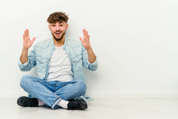 若いモロッコ人男性の床の上に白い背景に座って快適な驚きを受け、興奮し、手を上げる. - 写真・画像