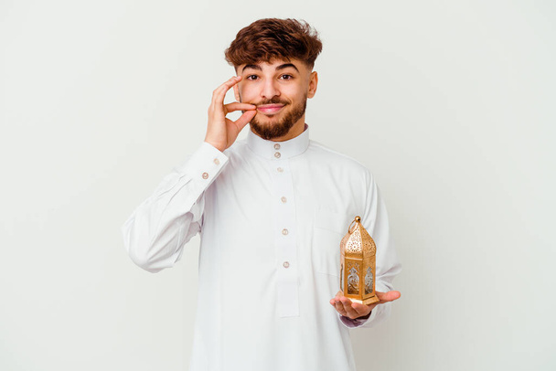 Młody Marokańczyk w typowym arabskim ubraniu trzymający lampę ramadanu odizolowaną na białym tle z palcami na ustach trzymającymi tajemnicę. - Zdjęcie, obraz