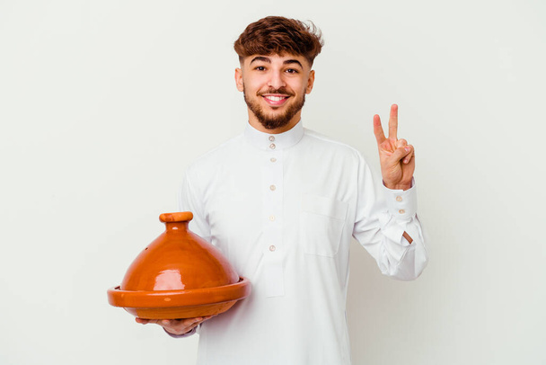 Young Moroccan man wearing the typical arabic costume holding a tajine isolated on white background showing number two with fingers. - Photo, Image