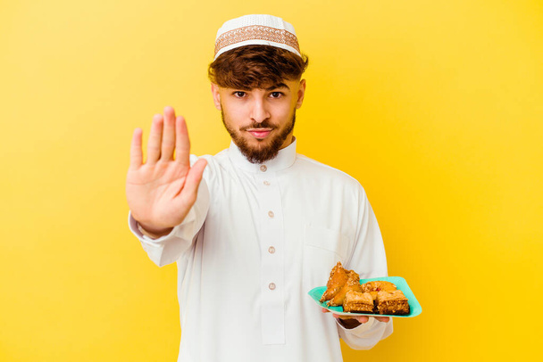 Joven marroquí vestido con el típico traje árabe comiendo dulces árabes aislados sobre fondo amarillo de pie con la mano extendida mostrando señal de stop, impidiendo. - Foto, imagen