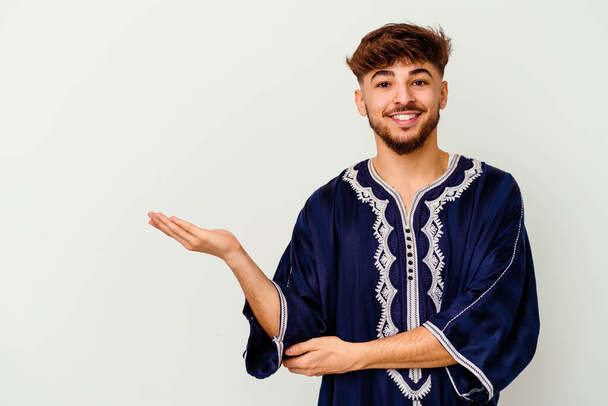 Jovem marroquino isolado no fundo branco mostrando um espaço de cópia em uma palma e segurando outra mão na cintura. - Foto, Imagem