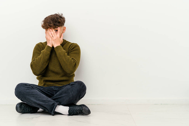 Junger marokkanischer Mann, der isoliert auf weißem Hintergrund auf dem Boden sitzt, blinzelt verängstigt und nervös durch die Finger. - Foto, Bild