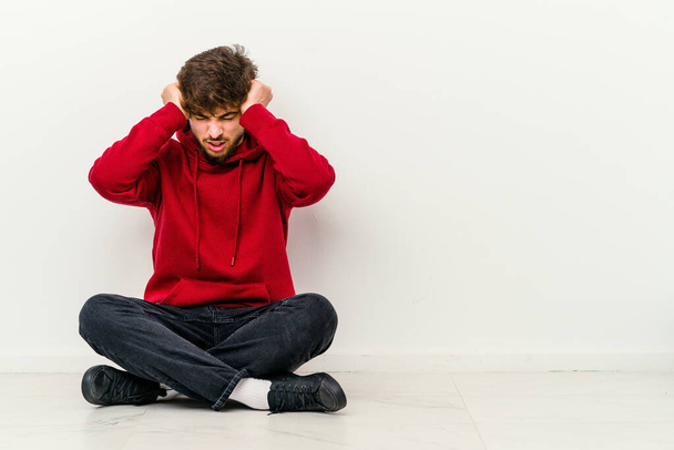 Young Moroccan man sitting on the floor isolated on white background covering ears with hands trying not to hear too loud sound. - Photo, Image