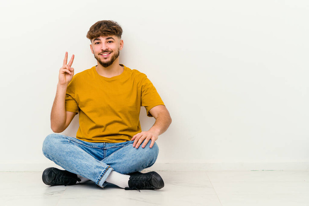 Молодой марокканский мужчина сидит на полу изолированы на белом фоне, показывая номер два с пальцами. - Фото, изображение