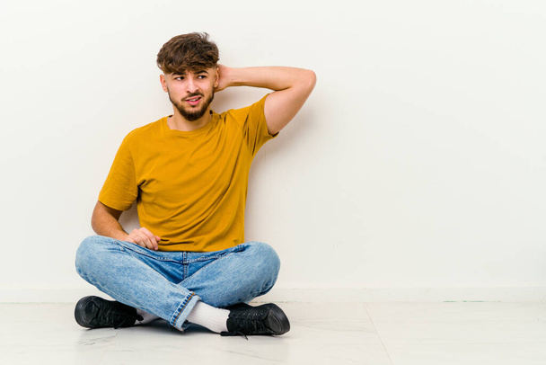 Faslı genç bir adam beyaz arka planda izole bir şekilde yerde oturuyor yorgun ve uykulu bir şekilde ellerini başının üstünde tutuyor.. - Fotoğraf, Görsel