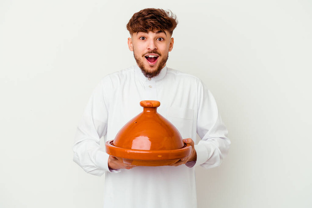 Young Moroccan man wearing the typical arabic costume holding a tajine isolated on white background receiving a pleasant surprise, excited and raising hands. - Photo, Image
