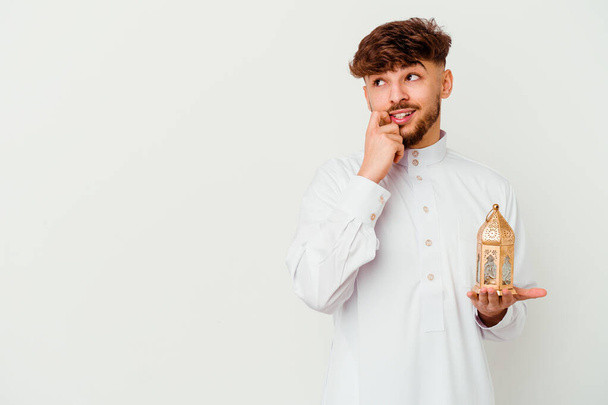 Junger marokkanischer Mann in typischer arabischer Kleidung hält eine Ramadan-Lampe auf weißem Hintergrund und denkt entspannt an etwas, das einen Kopierraum betrachtet. - Foto, Bild