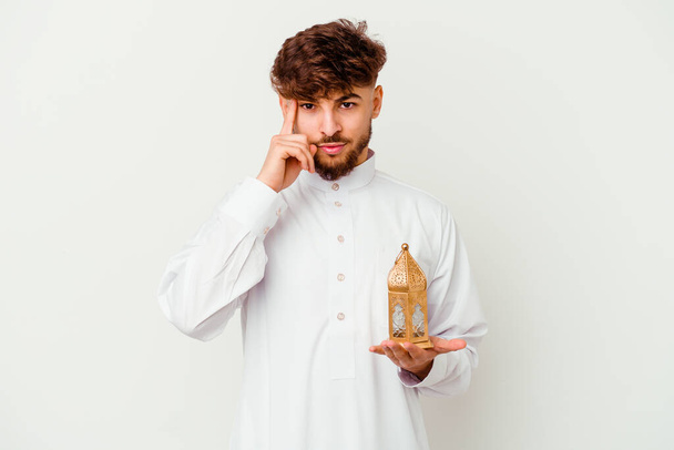 Νεαρός Μαροκινός άνδρας φορώντας ένα τυπικό arab ρούχα κρατώντας μια λάμπα ραμαντάν απομονώνονται σε λευκό φόντο δείχνοντας ναό με το δάχτυλο, σκέψης, επικεντρώθηκε σε μια εργασία. - Φωτογραφία, εικόνα