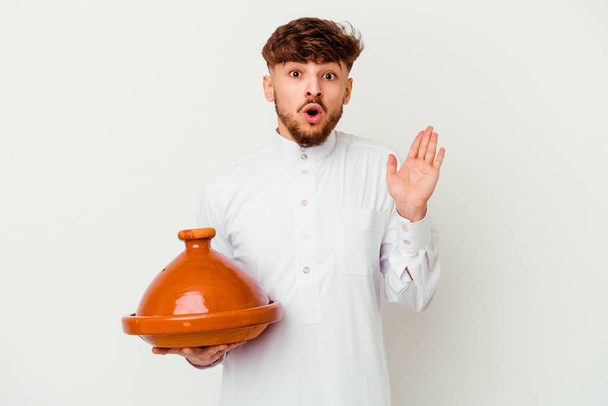 Joven marroquí vestido con el típico traje árabe sosteniendo un tajine aislado sobre fondo blanco sorprendido y sorprendido. - Foto, Imagen