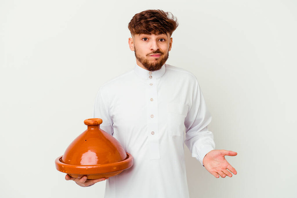 Młody Marokańczyk noszący typowy arabski kostium trzymający tajine na białym tle zdezorientowany, czuje się wątpliwy i niepewny. - Zdjęcie, obraz