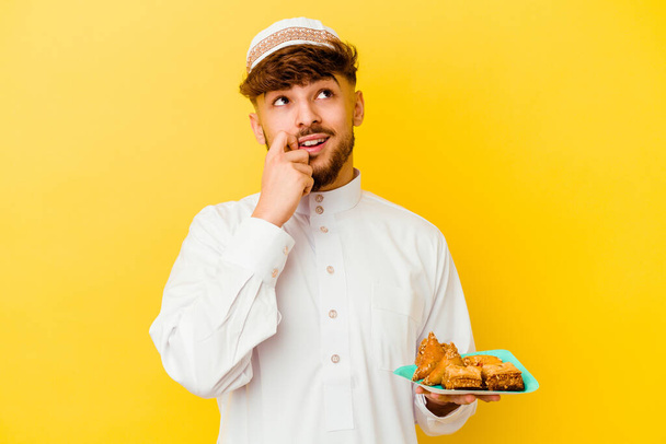 Junger marokkanischer Mann im typischen arabischen Kostüm isst arabische Süßigkeiten isoliert auf gelbem Hintergrund und denkt entspannt an etwas, das einen Kopierraum betrachtet. - Foto, Bild