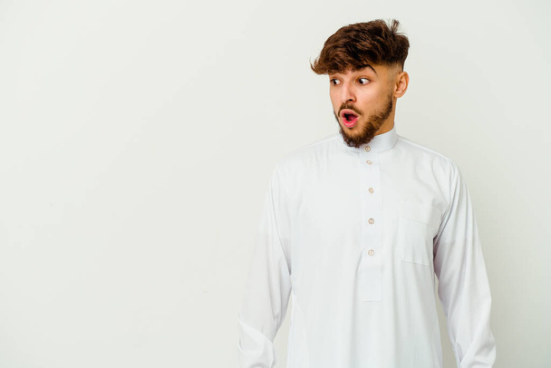 Junger marokkanischer Mann, der eine typisch arabische Kleidung auf weißem Hintergrund trägt, ist schockiert, weil sie etwas gesehen hat. - Foto, Bild