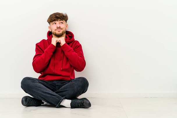 Young Moroccan man sitting on the floor isolated on white background keeps hands under chin, is looking happily aside. - Photo, Image