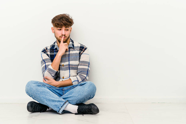 Jeune homme marocain assis sur le sol isolé sur fond blanc regardant de côté avec une expression douteuse et sceptique. - Photo, image