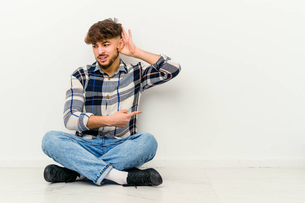 Joven hombre marroquí sentado en el suelo aislado sobre fondo blanco tratando de escuchar un chisme. - Foto, imagen