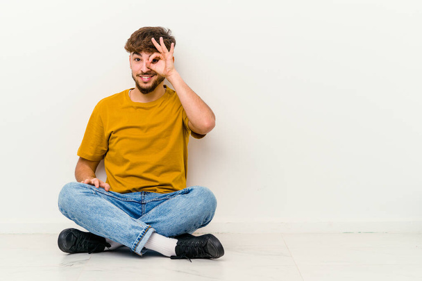 Young Moroccan man sitting on the floor isolated on white background excited keeping ok gesture on eye. - Φωτογραφία, εικόνα