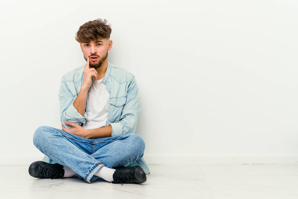 Joven hombre marroquí sentado en el suelo aislado sobre fondo blanco mirando hacia los lados con expresión dudosa y escéptica. - Foto, Imagen