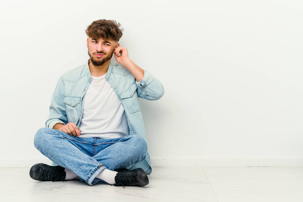 Mladý Maročan sedí na podlaze izolovaný na bílém pozadí a zakrývá uši prsty, vystresovaný a zoufalý hlasitým okolím. - Fotografie, Obrázek