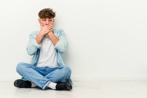Jovem marroquino sentado no chão isolado no fundo branco pensativo olhando para um espaço de cópia cobrindo boca com a mão. - Foto, Imagem