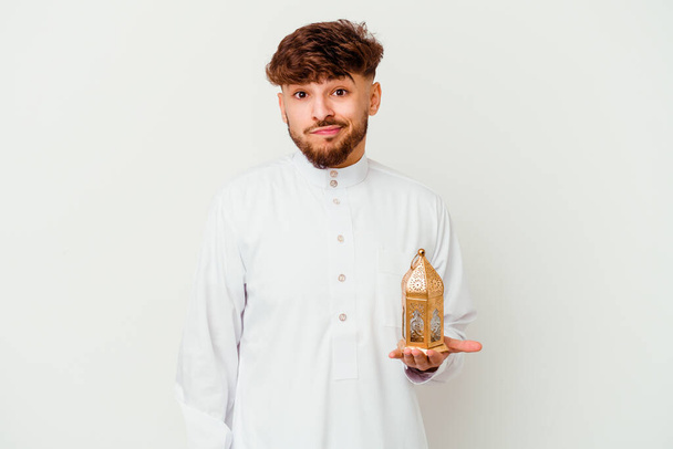Giovane marocchino indossa un tipico abiti arabi in possesso di una lampada ramadan isolato su sfondo bianco confuso, si sente dubbioso e incerto. - Foto, immagini
