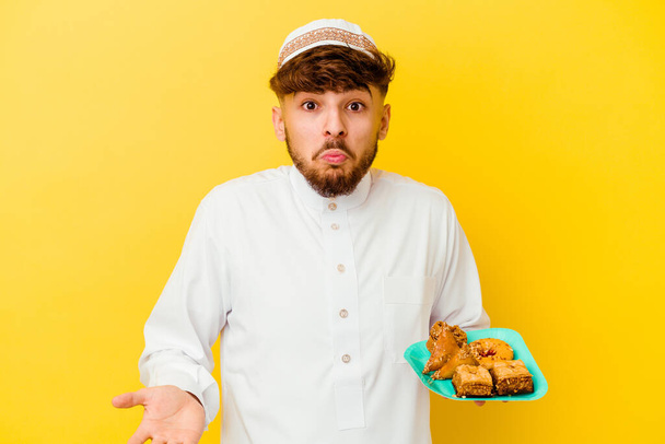 Νεαρός Μαροκινός άνδρας φορώντας το τυπικό αραβικό κοστούμι τρώει αραβικά γλυκά που απομονώνονται σε κίτρινο φόντο ανασηκώνει τους ώμους και ανοιχτά μάτια σύγχυση. - Φωτογραφία, εικόνα