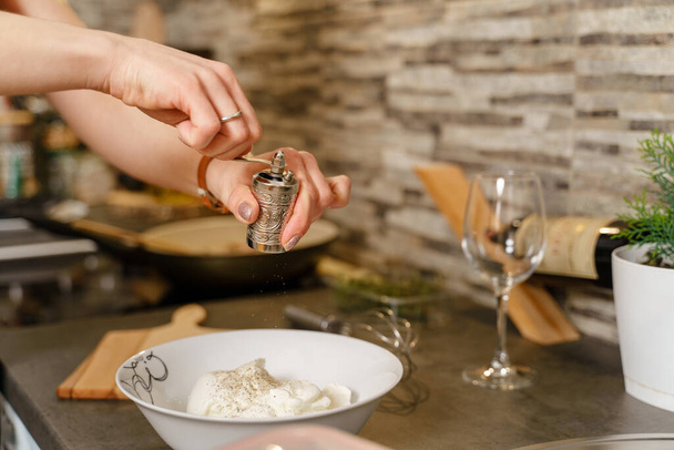Adding spices to the meal close up on hands of unknown caucasian woman holding pepper grinder above plate with cream while cooking and preparing sauce food at home in the kitchen - Photo, image