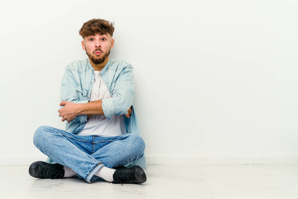 Young Moroccan man sitting on the floor isolated on white background shrugs shoulders and open eyes confused. - Photo, Image