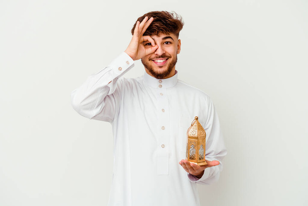 Νεαρός Μαροκινός άνδρας φορώντας ένα τυπικό arab ρούχα κρατώντας μια λάμπα ραμαντάν απομονώνονται σε λευκό φόντο ενθουσιασμένος κρατώντας ok χειρονομία στο μάτι. - Φωτογραφία, εικόνα