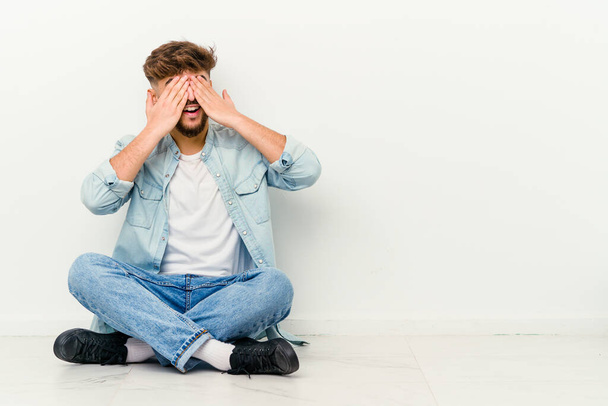 Jovem marroquino sentado no chão isolado no fundo branco com medo de cobrir os olhos com as mãos. - Foto, Imagem