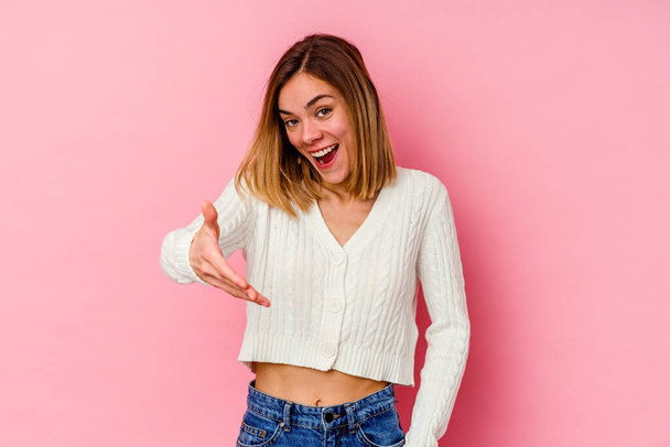 Young caucasian woman isolated on pink background stretching hand at camera in greeting gesture. - Foto, afbeelding