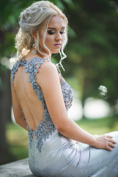 A closeup shot of a shy blond woman sitting outdoors in a silver dress with an open back - Φωτογραφία, εικόνα