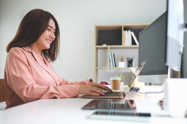 Uśmiechnięta młoda kobieta pracująca z laptopem w nowoczesnym biurze. - Zdjęcie, obraz