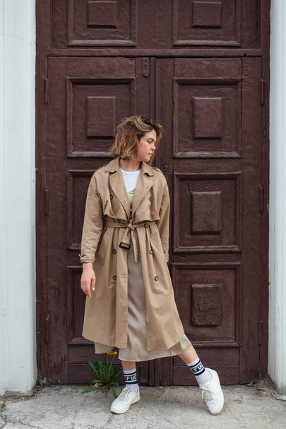 Young smiling millennial woman with wild hair dressed in an autumn coat posing near the door of an old building. - Foto, imagen