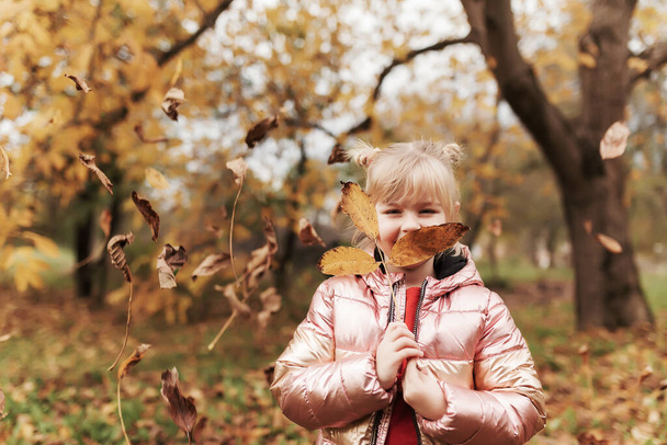 Malá blondýnka si hraje se žlutými podzimními listy na zahradě, usmívá se, baví - Fotografie, Obrázek