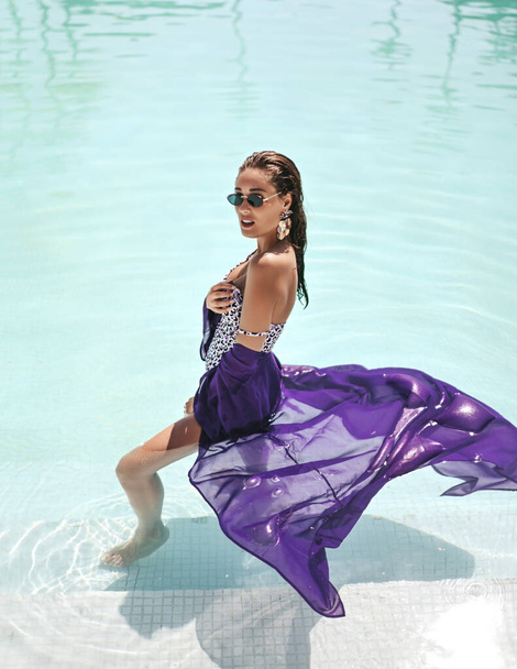 Rich sexy woman with big breast in white leopard print bikini and purple pareo stands sideways in pool during vacation - Φωτογραφία, εικόνα